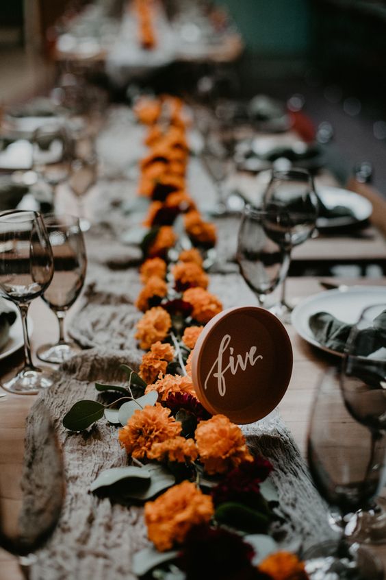 a chic and simple wedding tablescape with a grey linen table runner and marigolds and greenery, a table number and neutral porcelain
