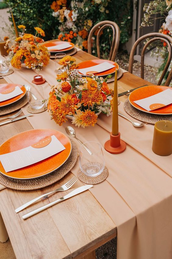 A chic mid century modern wedding tablescape with a blush table runner, orange and rust blooms, orange candles and porcelain, woven chargers and woven coasters