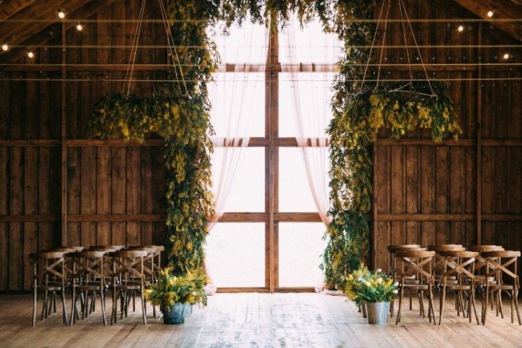  a rustic wedding ceremony space with a double-height window covered with mimosa as an arch, chandeliers and arrangements along the aisle