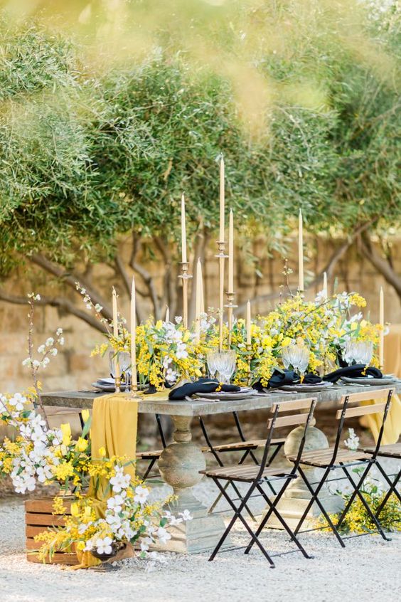 a beautiful spring wedding tablescape with a table of concrete, mimosa and cherry blossom, a yellow runner, black napkins and tall and thin candles