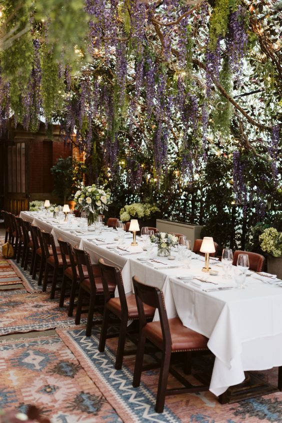 a cozy and dreamy wedding reception space with wisteria hanging down and greenery, a long table and table lamps plus leather chairs