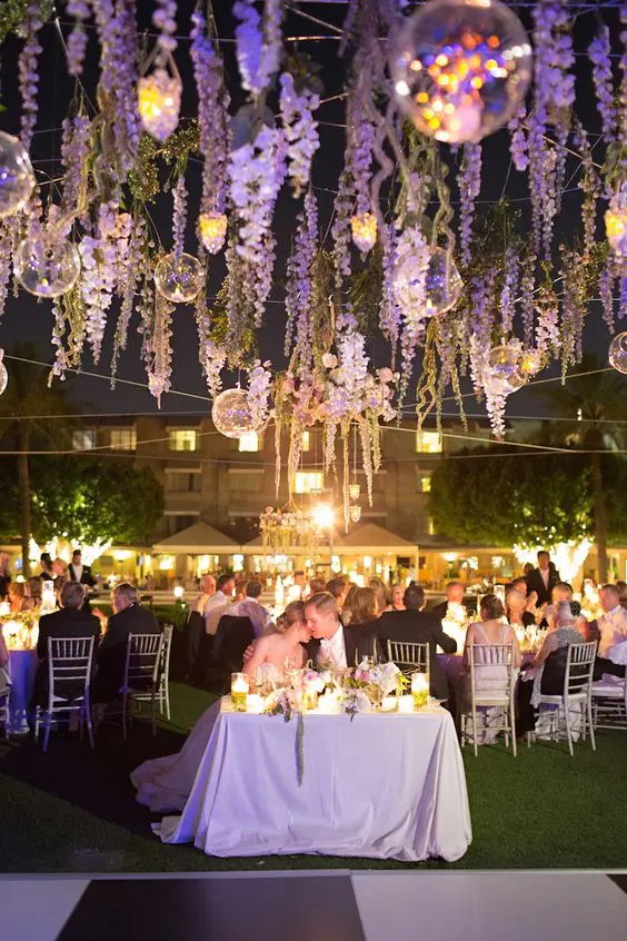 a cozy outdoor wedding ceremony space with wisteria hanging down, some candle lanterns and lights all over the space