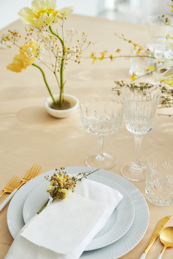 a delicate spring wedding tablescape with grey plates, mimosa and yellow poppies, chic modern cutlery and crystal glasses