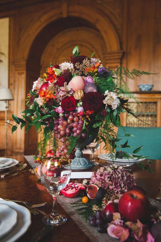 a super bold and lush fall wedding centerpiece of white, orange and burgundy blooms, greenery, grapes and pears and matching stuff on the table