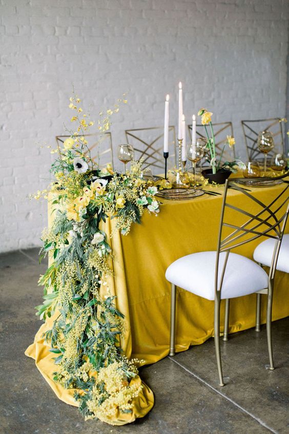 a lovely spring wedding table setting with a yellow tablecloth, mimosa, anemones and ranucnulus, daffodils and tall and thin candles