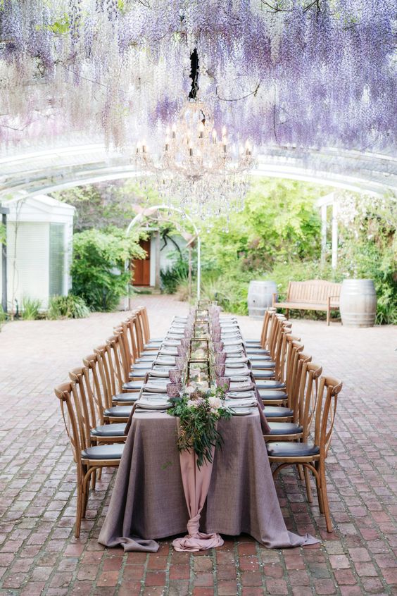 a lovely wedding reception space with wisteria naturally growing over it, with a table and a lilac tablecloth, purple glasses and greenery