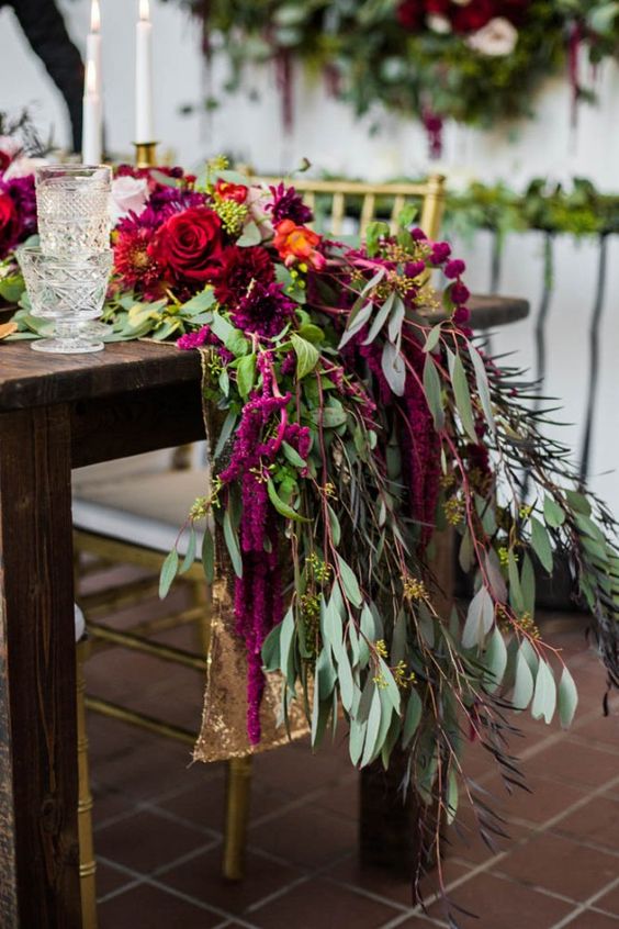 a lush wedding table runner of greenery, red roses and dahlias, berries and amaranthus for a bold fall wedding