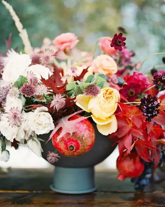 an adorable fall wedding centerpiece of a grey vase, yellow, pink and deep red blooms, fall leaves and pomegranates that help to embrace the fall