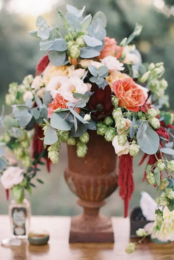 a soft wedding centerpiece of blush and red orange blooms plus pale greenery in a vintage urn