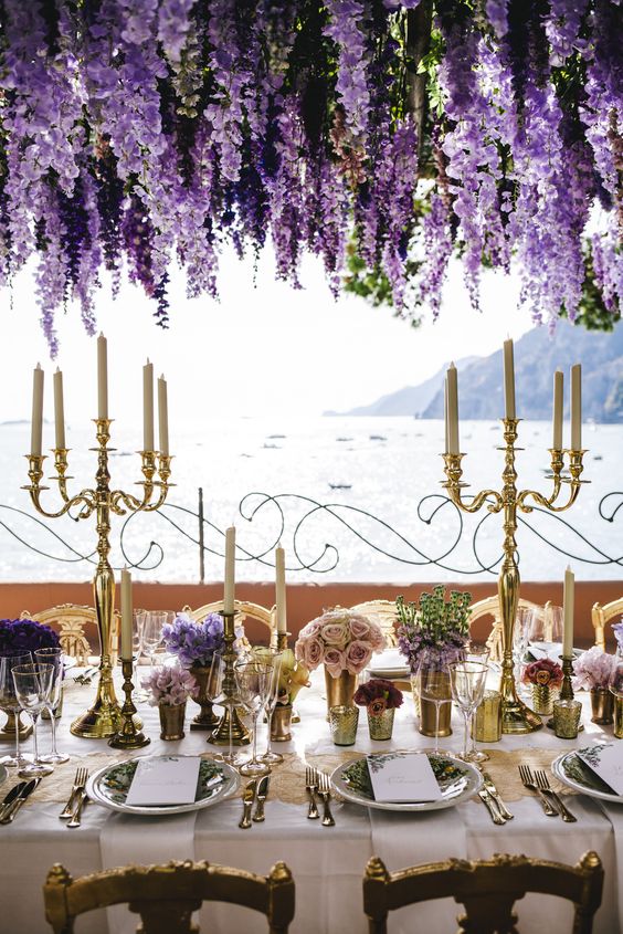 a luxurious wedding reception space with a lake view, a wisteria overhead installation, lilac, purple, blush blooms in pots and candelabras