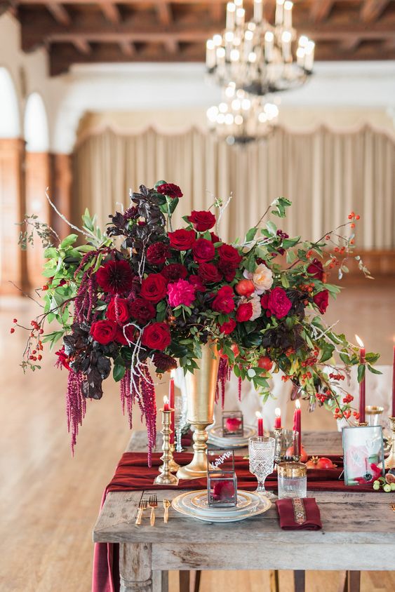 a vibrant fall wedding centerpiece of red and blush roses, burgundy dahlias, dark foliage and greenery and amaranthus