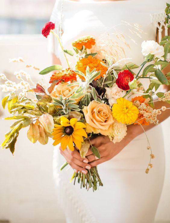 a bright fall wedding bouquet of sunflowers, roses, marigolds, ranunculus, greenery and grasses for a fall wedding