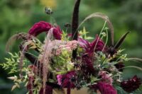 a sophisticated moody wedding centerpiece of greenery, burgundy dahlias and anemones, feathers, grasses and amaranthus