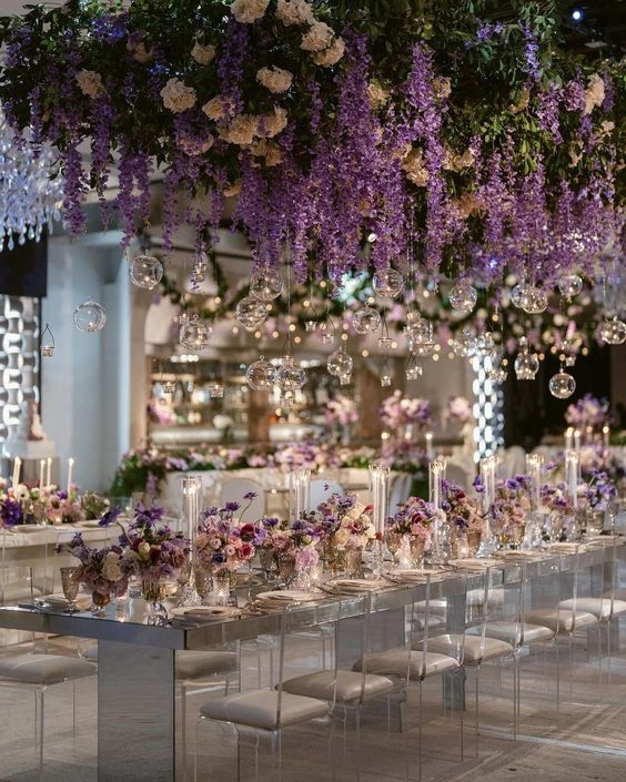 a refined and glam wedding reception space with a mirrored table and clear chairs, an oversized overhead wedding installation of white blooms and wisteria plus bulbs