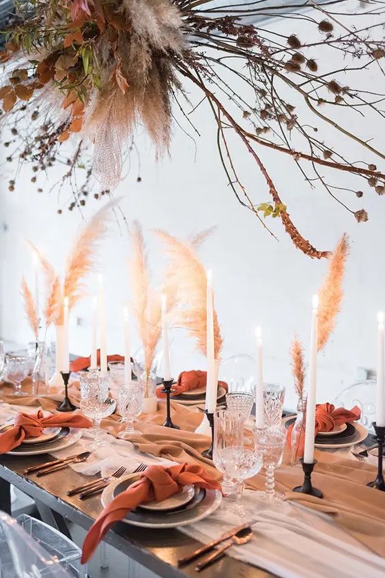 a boho wedding tablescape with a pastel runner, rust napkins, grasses, candles and a large installation with rust leaves, branches and pampas grass