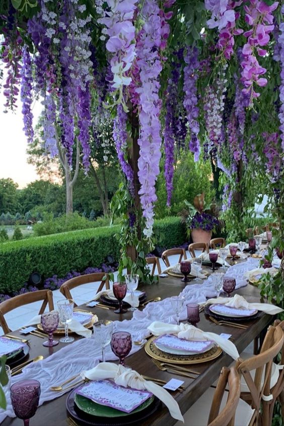 a super lush wedding reception space with an overhead floral installation with wisteria, a lilac table runner and purple glasses