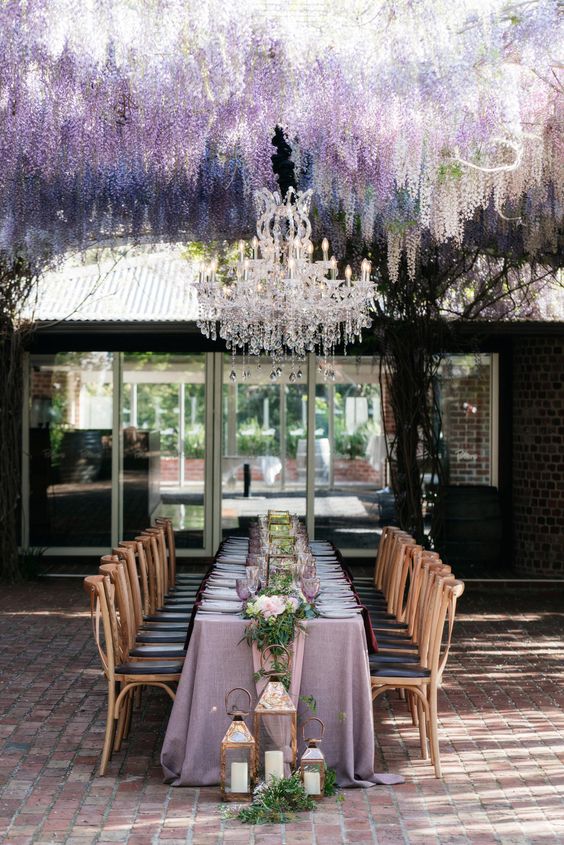 an outdoor wedding reception space with wisteria hanging over the table, a large crystal chandelier, a refined tablescape with a lilac tablecloth