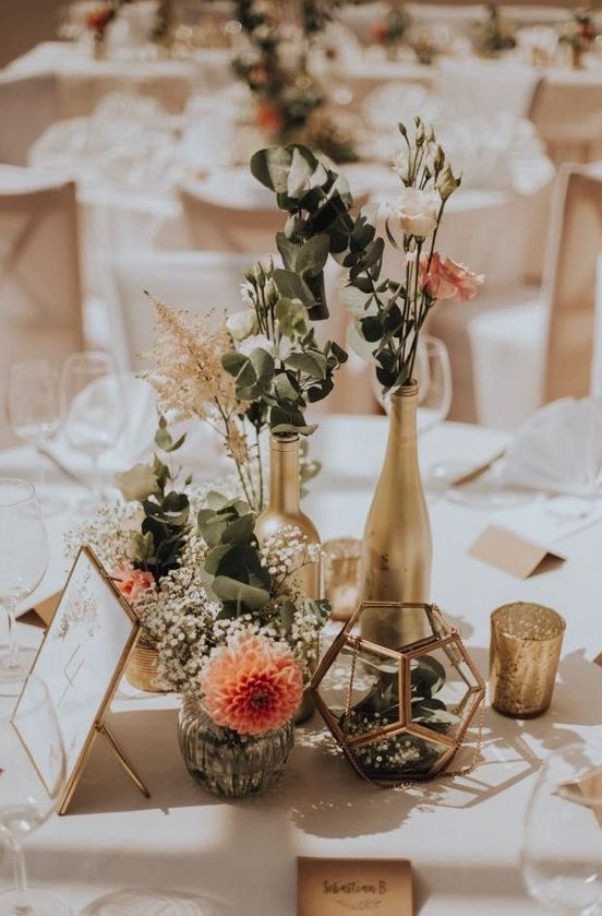 a cluster fall wedding centerpiece of gilded bottles and candleholders, greenery, white and pink roses, candles and baby's breath