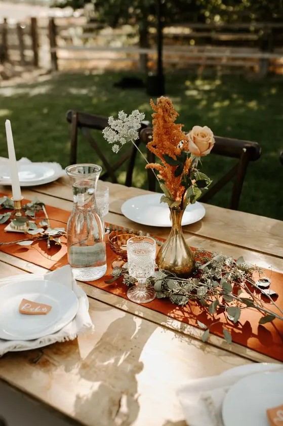 a cool and simple fall boho wedding centerpiece of a gilded vase, a blush rose, white and rust blooms, greenery on the table and a rust-colored table runner