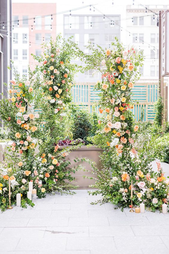 a lush wedding arch done with greenery, blush and pink blooms, some marigolds, candles of various kinds