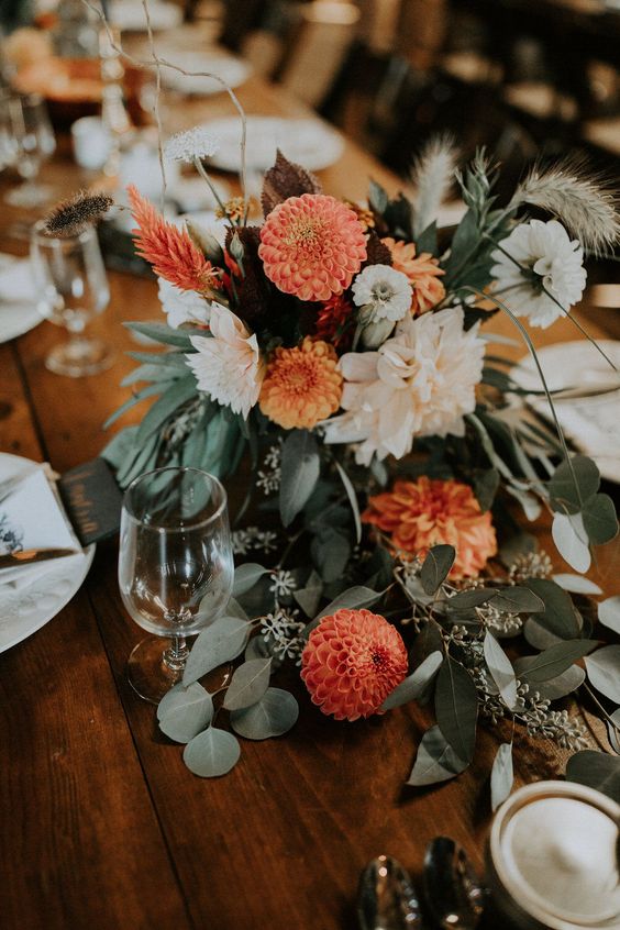 a pretty fall wedding centerpiece of white and blush dahlias, orange mums, greenery and grasses is a lovely idea for a boho wedding