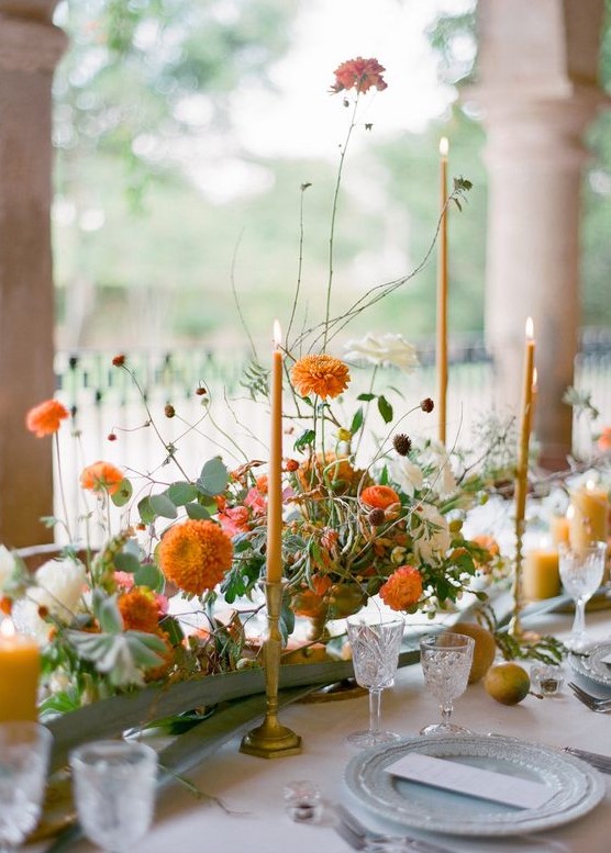 a pretty fall wedding centerpiece of white and orange blooms, greenery and orange candles is refined and chic