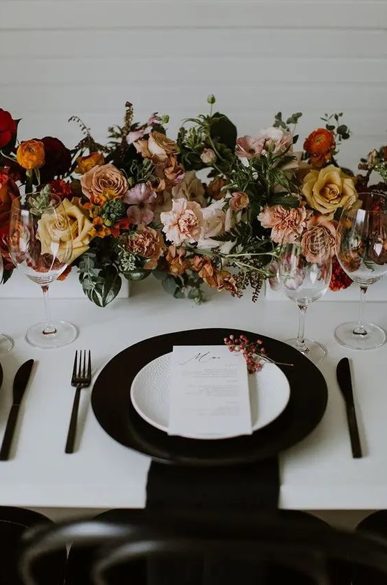 a stylish modern fall wedding tablescape with a black charger, black cutlery, lovely blush, peachy, rust and yellow blooms and greenery
