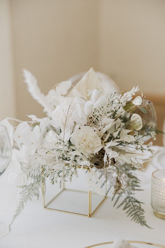 a beautiful white wedding centerpiece on a gold frame, with white carnations, orchids, lunaria, greenery and pampas grass is chic