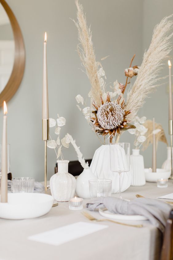 a boho cluster wedding centerpiece of white vases, pampas grass, cotton, king protea, lunaria and tall and thin candles is wow