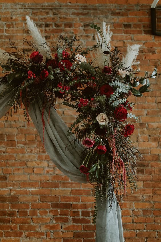 a boho wedding arch with grey fabric, greenery, white and burgundy blooms, pampas grass and a bit of amaranthus