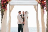 a bold tropical wedding arch covered with bougainvillea, with bright roses and ranunculus and greenery is amazing for a beach wedding