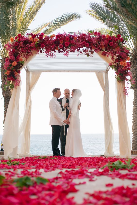 a bold tropical wedding arch covered with bougainvillea, with bright roses and ranunculus and greenery is amazing for a beach wedding
