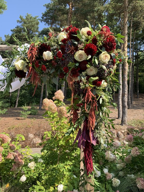 a bold wedding arch with greenery, white roses, burgundy dahlias, amaranthus and dark foliage is a cool idea for the fall