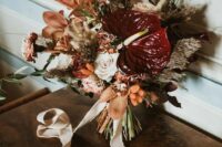 a catchy wedding bouquet of burgundy anthurium, rust lilies and dried and fresh blooms and grasses plus ribbons
