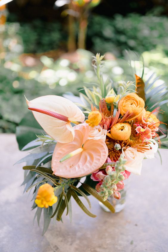 a catchy wedding centerpiece of yellow ranunculus, blush anthuriums, some bold tropical fillers and greenery for a tropical wedding