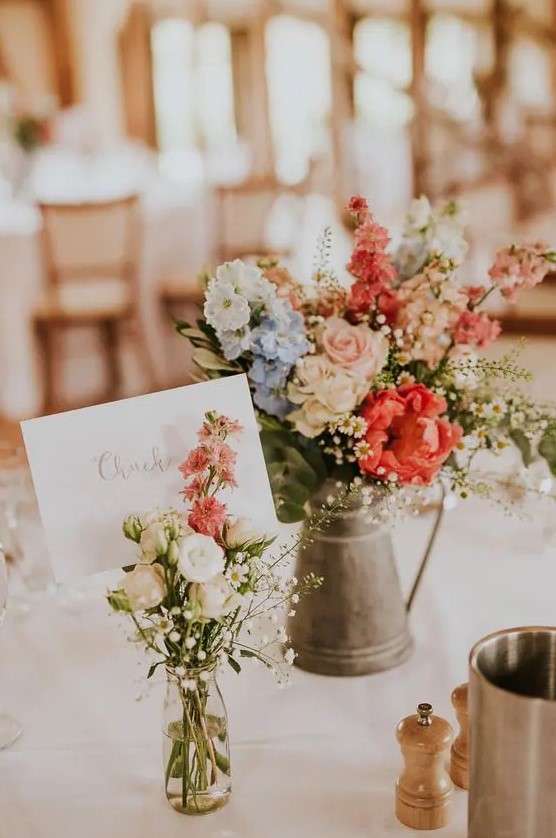 a cluster rustic wedding centerpiece of a metal jug with bright blooms and greenery and a jar with a neutral floral arrangement