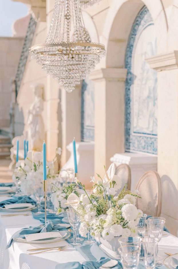 a coastal wedding tablescape with a blue table runner, napkins and candles, white florals with anthurium and gold cutlery