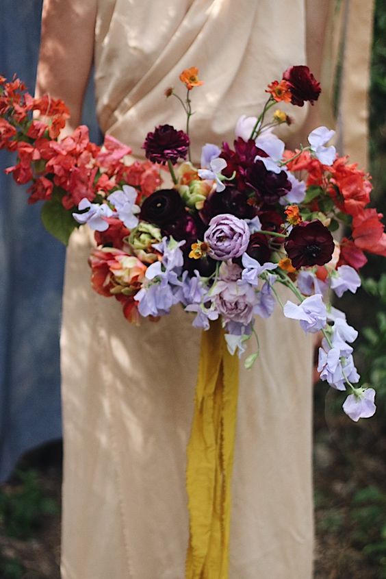 a colorful and contrasting wedding bouquet of red, deep purple, lilac blooms including ranunculus and swete peas is wow