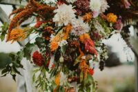 a colorful fall wedding arch with white and blush dahlias, red mums, greenery, grasses and amaranthus is a cool idea for a fall wedding