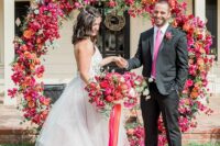 a colorful round wedding arch with bougainvillea, roses, citrus, orange blooms and greenery and a matching bridal bouquet
