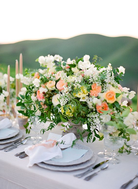 a colorful summer wedding centerpiece of lush greenery and fillers, white and orange ranunculus and white sweet peas