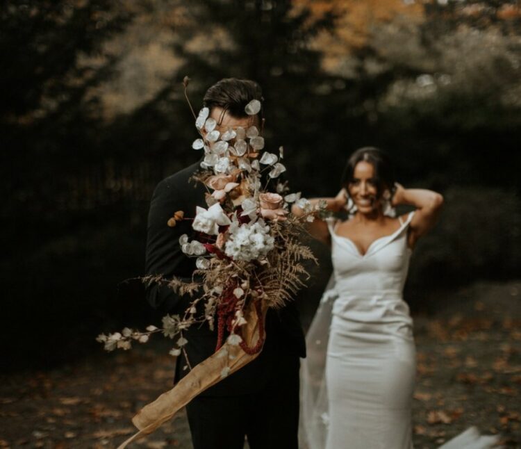 a creative boho wedding bouquet of white and blush roses, lunaria, dried leaves, greenery and amaranthus and long ribbon for a fall wedding