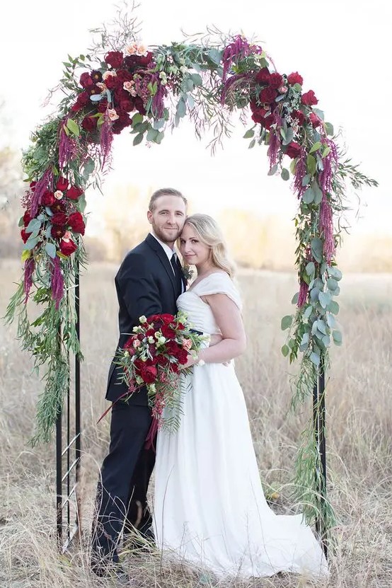 a delicate fall wedding arch decorated with peachy blooms, burgundy ones, deep red blooms and lots of greenery