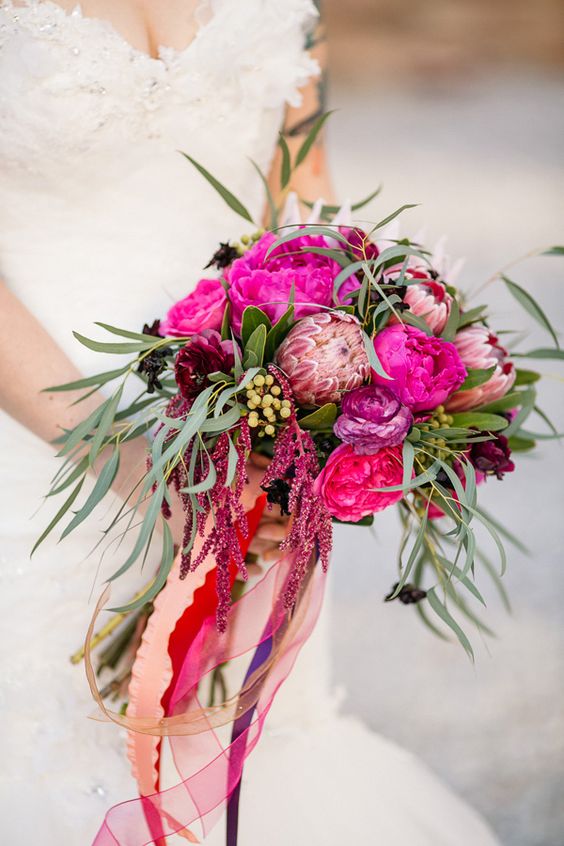 a jewel-tone wedding bouquet of hot pink and fuchsia blooms, king protea, greenery, berries and amaranthus plus long colorful ribbons