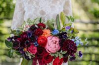 a jewel-tone wedding bouquet of red roses, purple and burgundy blooms, orange and blue ones, greenery and amaranthus for a colorful wedding