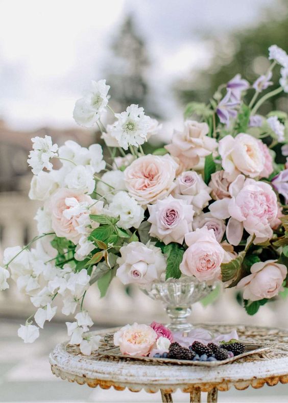 a lush and delicately colored wedding centerpiece of white sweet peas, blush peony roses, pale pink roses and orchids and geenery