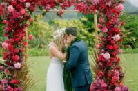 a lush tropical wedding arch covered with pink and red roses and bougainvillea, greenery for a bold-colored celebration