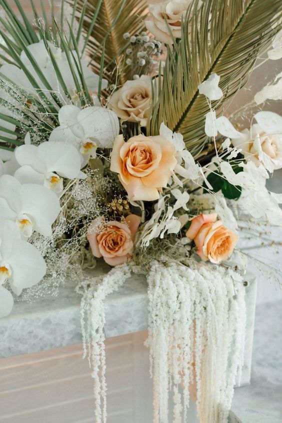 a lush tropical wedding arrangement of peachy roses, white orchids, amaranthus and usual and dried fronds plus lunaria is wow