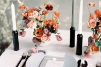 a modern bright wedding centerpiece with orange, blush and burgundy blooms in a white vase and tall and thin candles