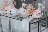 a modern wedding tablescape with a clear glass table and clear plates, pastel centerpieces of blush roses, anthuriums and lunarias